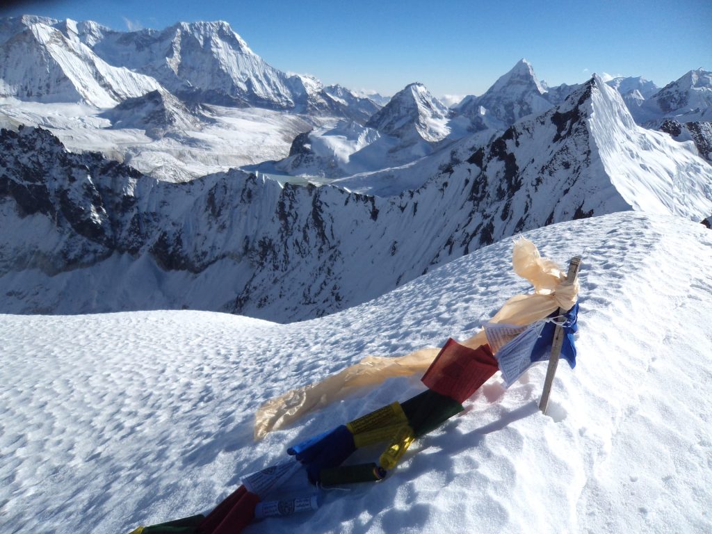 Brightly colored prayer flags fluttering in the wind on a high Himalayan mountain ridge, surrounded by vast sky and rugged peaks.