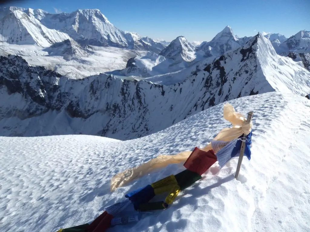 Brightly colored prayer flags fluttering in the wind on a high Himalayan mountain ridge, surrounded by vast sky and rugged peaks.