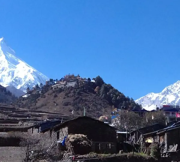 A small village nestled on top of a green hill surrounded by mountains along the Manaslu trekking route in Nepal.