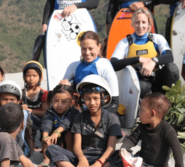 A group of children smiling and posing together with a rafting team, all wearing life jackets and helmets near a river in Nepal.